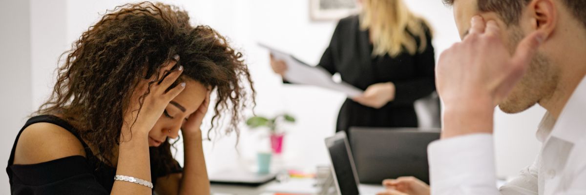 A woman holding her head in her hands in frustration at a desk while a colleague holds papers in the background.