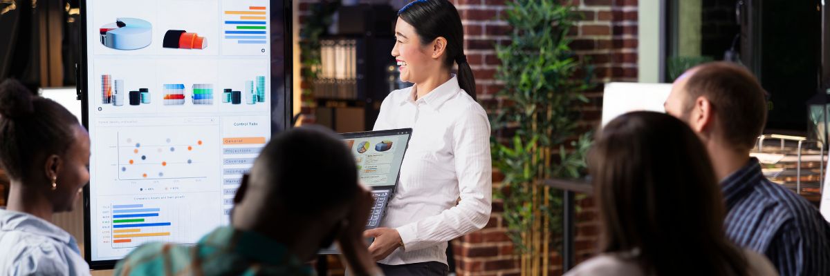 A smiling woman presents data charts on a large screen to a diverse group of colleagues in a modern office.
